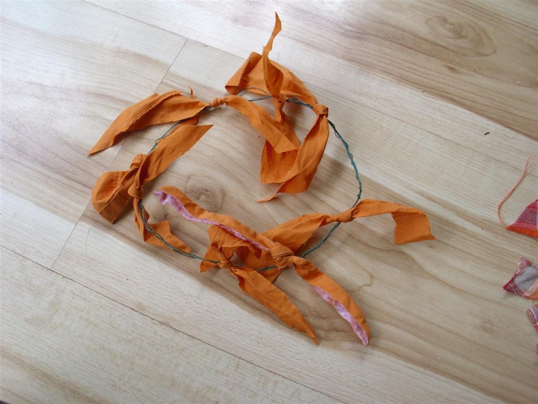 Orangefarbene Stoffstreifen, zu Knoten gebunden und kreisförmig auf einem Holzboden arrangiert. Dies illustriert eine handgefertigte, textile Verpackungslösung oder das Material in Vorbereitung dafür und unterstreicht, wie die Wiederverwendung von Stoffresten eine nachhaltige Alternative zu konventionellen Verpackungen darstellt.